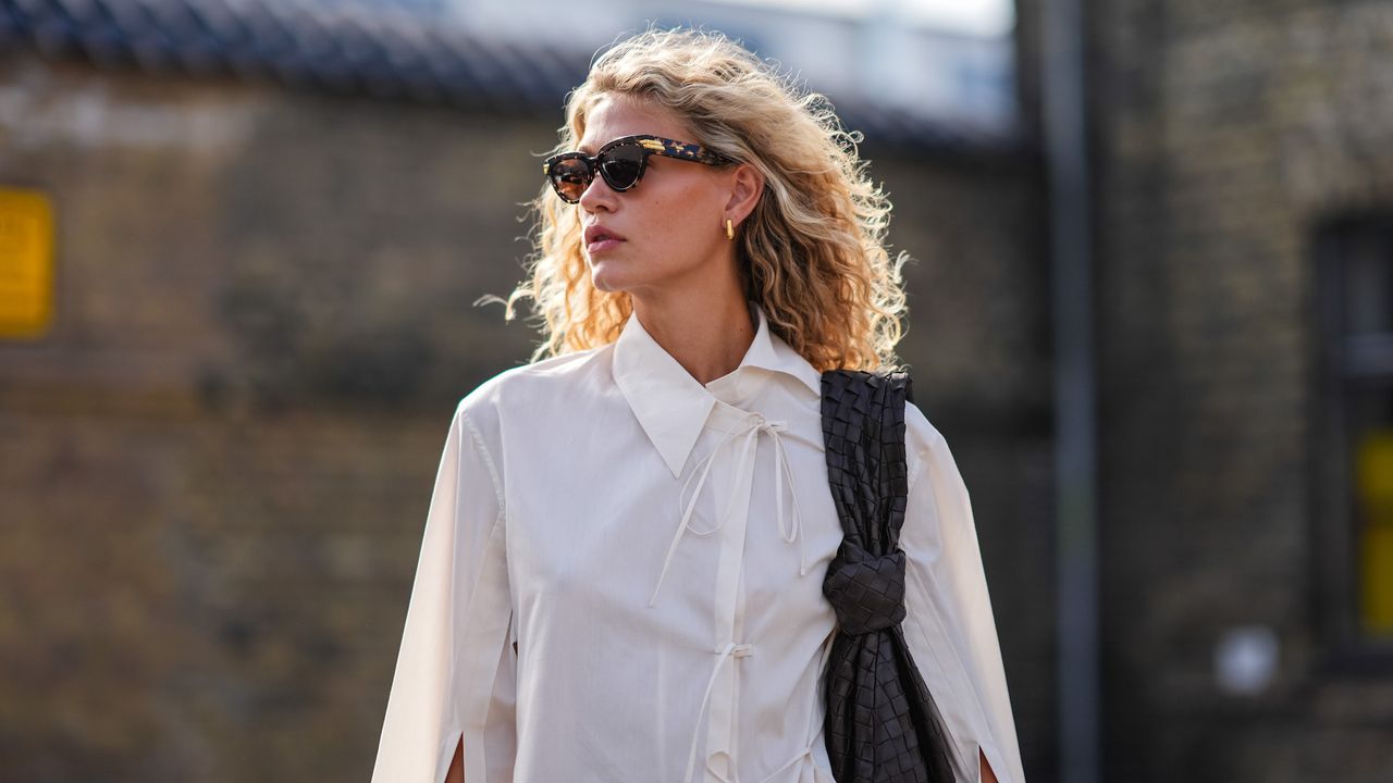 A young woman with wavy blonde air dried hair wears black sunglasses white button up shirt