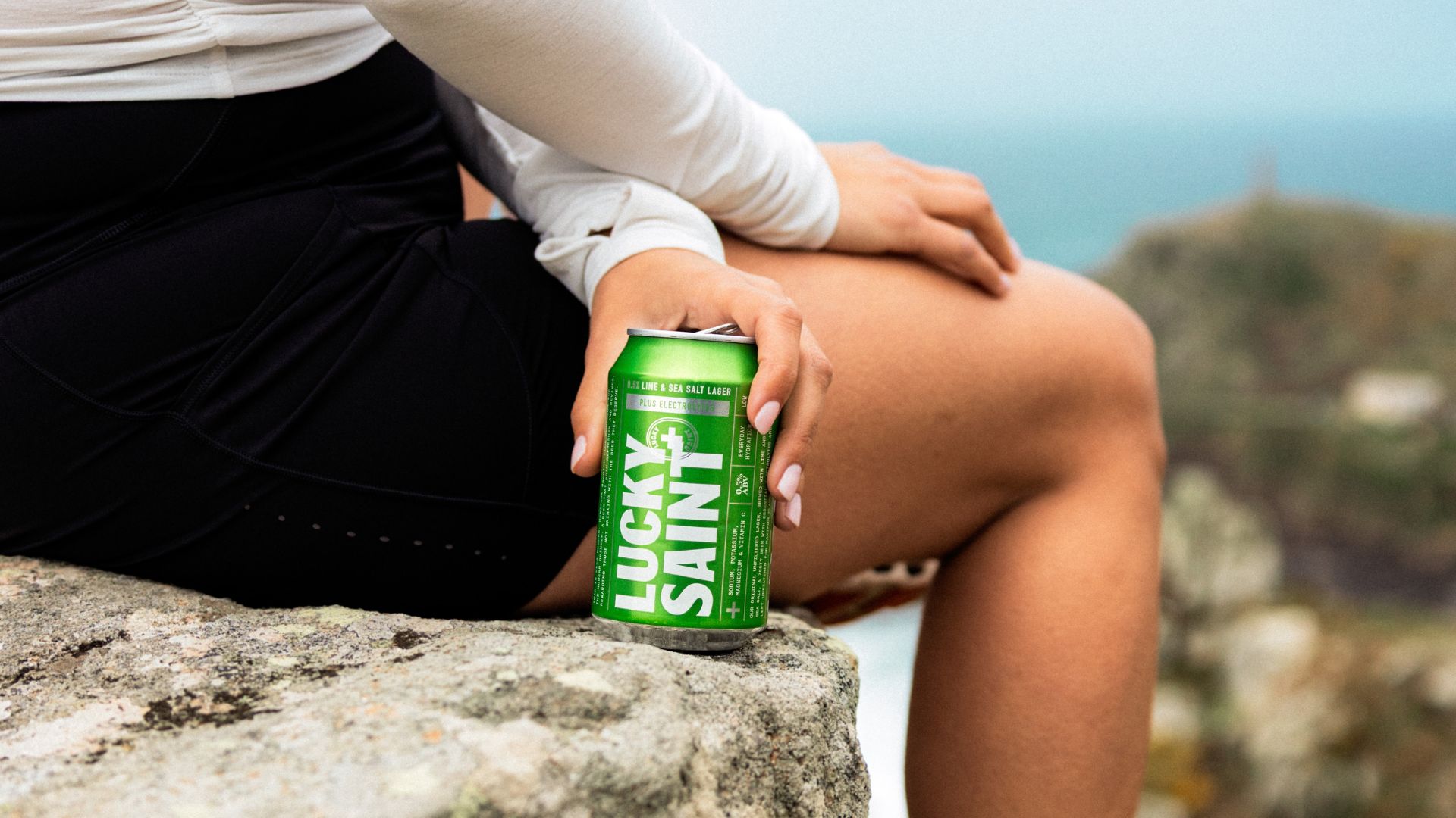 an image of the new lucky saint lime and sea salt larger in a green can being held by someone sitting on a rock mid-walk