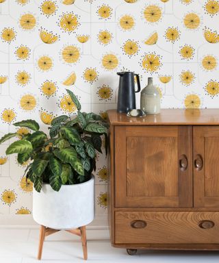 Fun, retro floral wallpaper in white and mustard colorway, behind potted plant on legs, and wood sideboard.