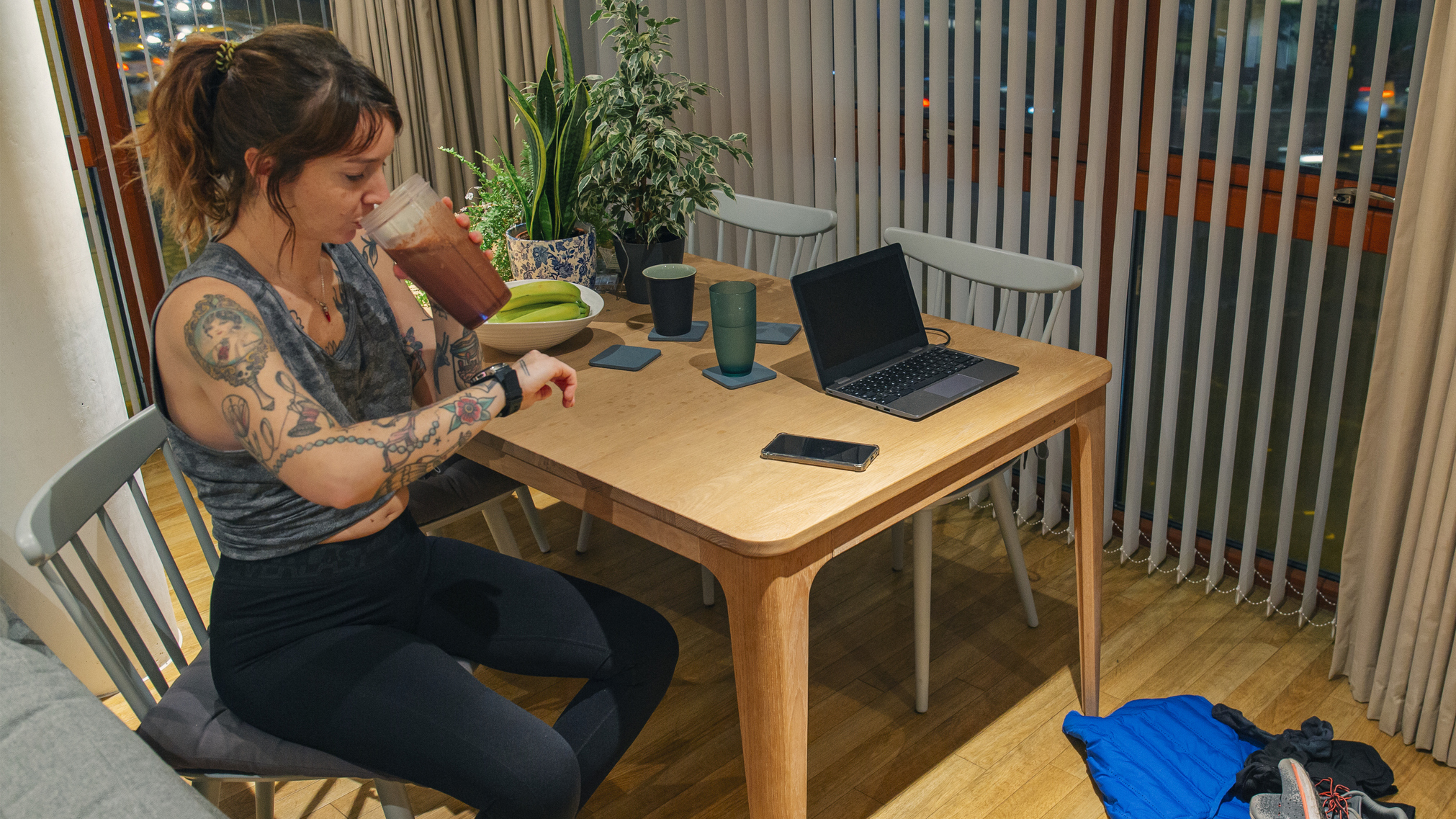 Woman drinks smoothie at a kitchen table and looks at her smartwatch