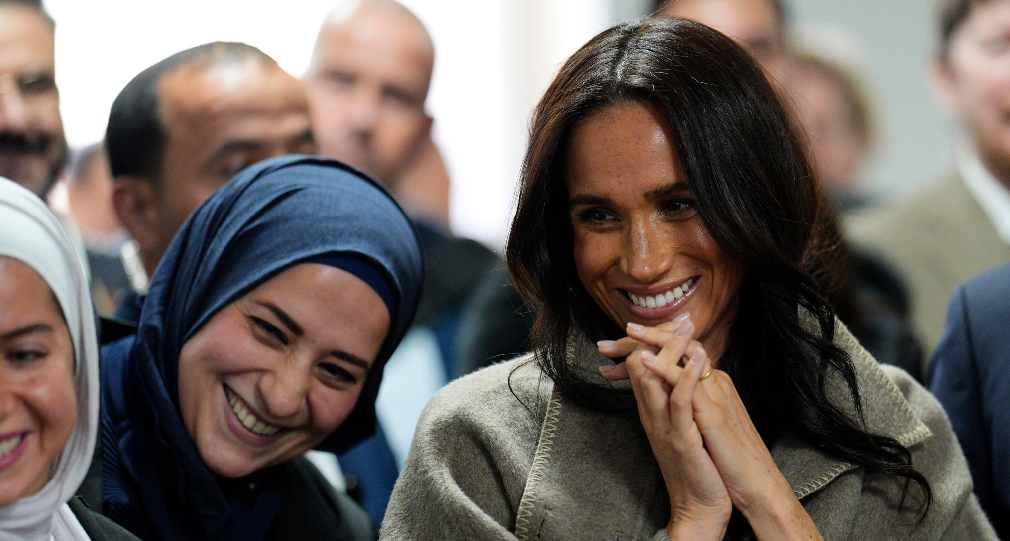 Meghan Markle clasping her hands and smiling in a taupe coat