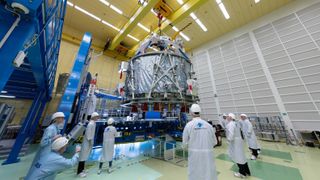 technicians in white suits surround a spacecraft on a hanging mechanism in a factory