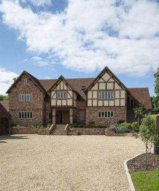 A brick and oak frame home with a large gravel driveway to the front of the image and a blue sky behind