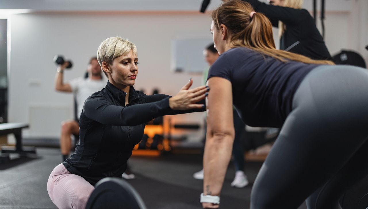 Personal trainer in a gym corrects the form of a woman gym-goer holding a barbell