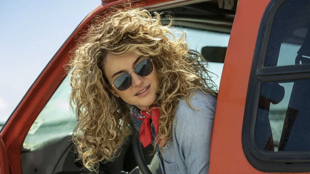 Actor Shailene Woodley (playing Gia), with frizzy hair and denim jacket, looks out of a red truck in the desert in a scene from TV drama Three Women