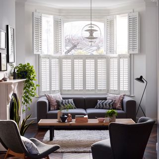 Wooden bay window shutters frame dark living room sofa and armchairs with house plants on mantle