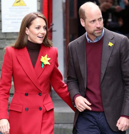 Kate Middleton wearing a red coat and yellow daffodil looking to the right walking outside next to Prince William, also looking at the same thing and wearing a daffodil on his blazer