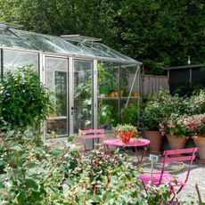 A close up of a flower garden with a greenhouse and pink table/seats 