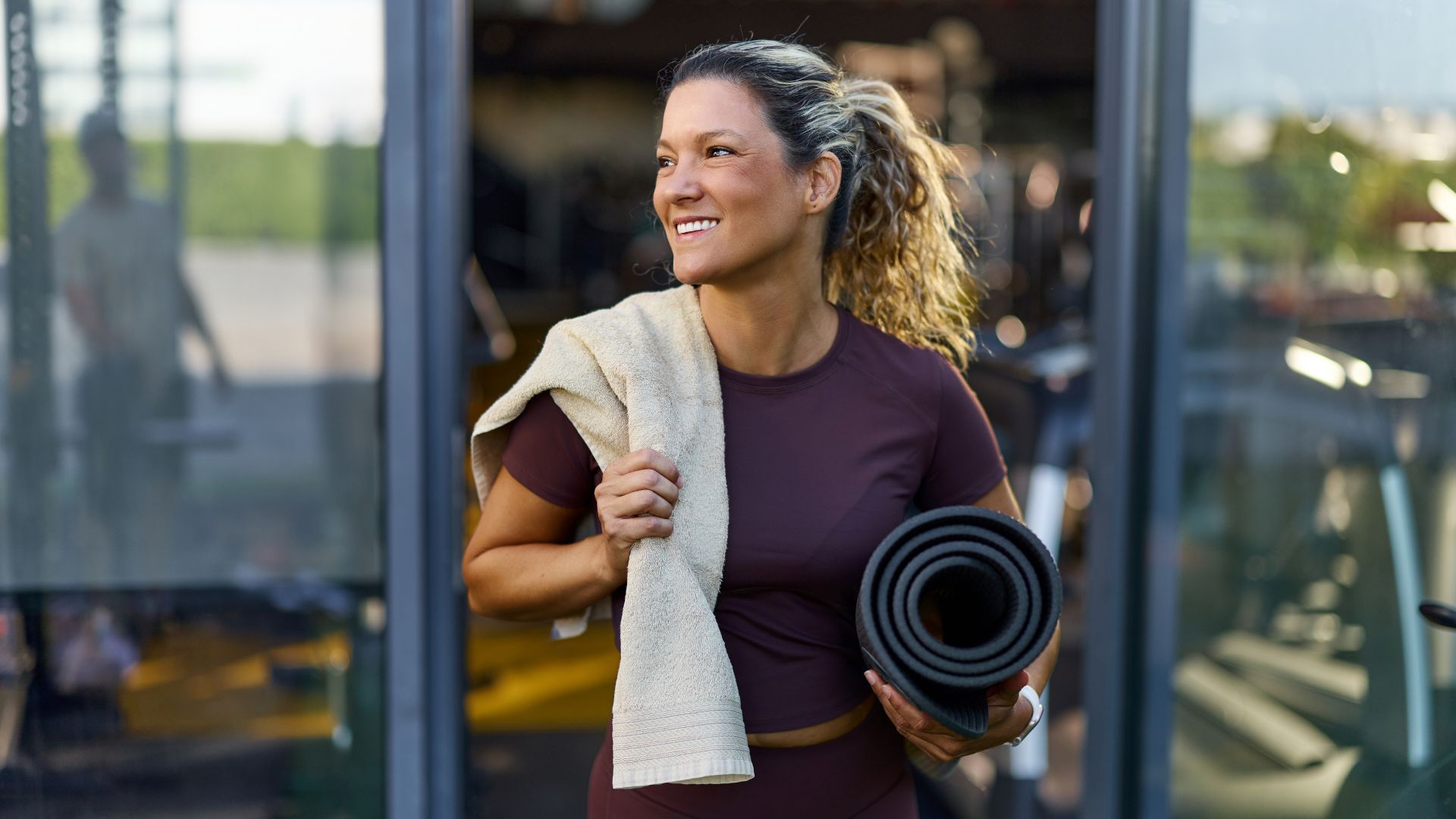 Woman setting up for boat pose, holding yoga mat and towel, coming out of studio smiling