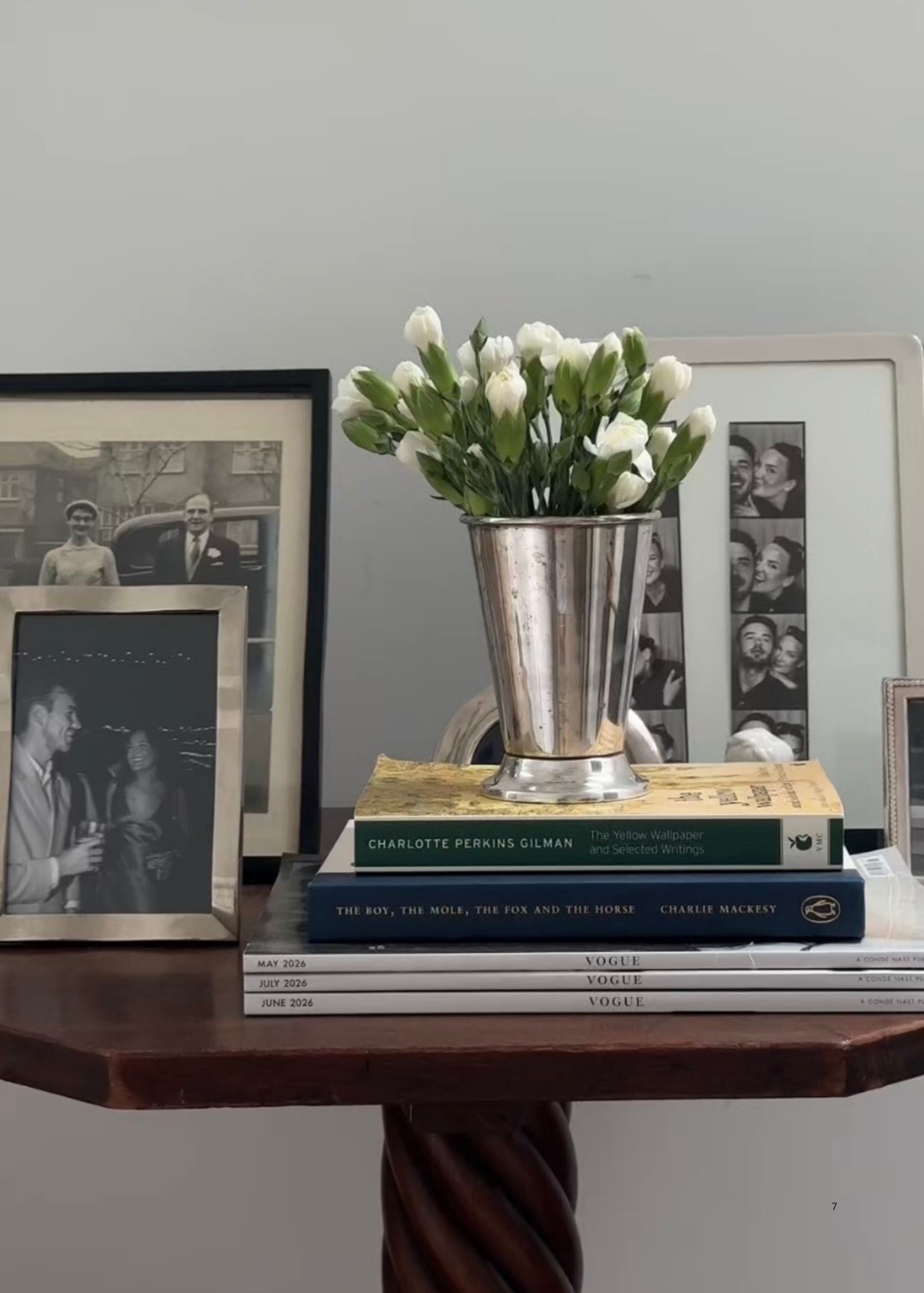 Close-up of a wooden table with a stack of Vogue magazines and books, old photographs in frames, and a silver vase with white tulips in it.