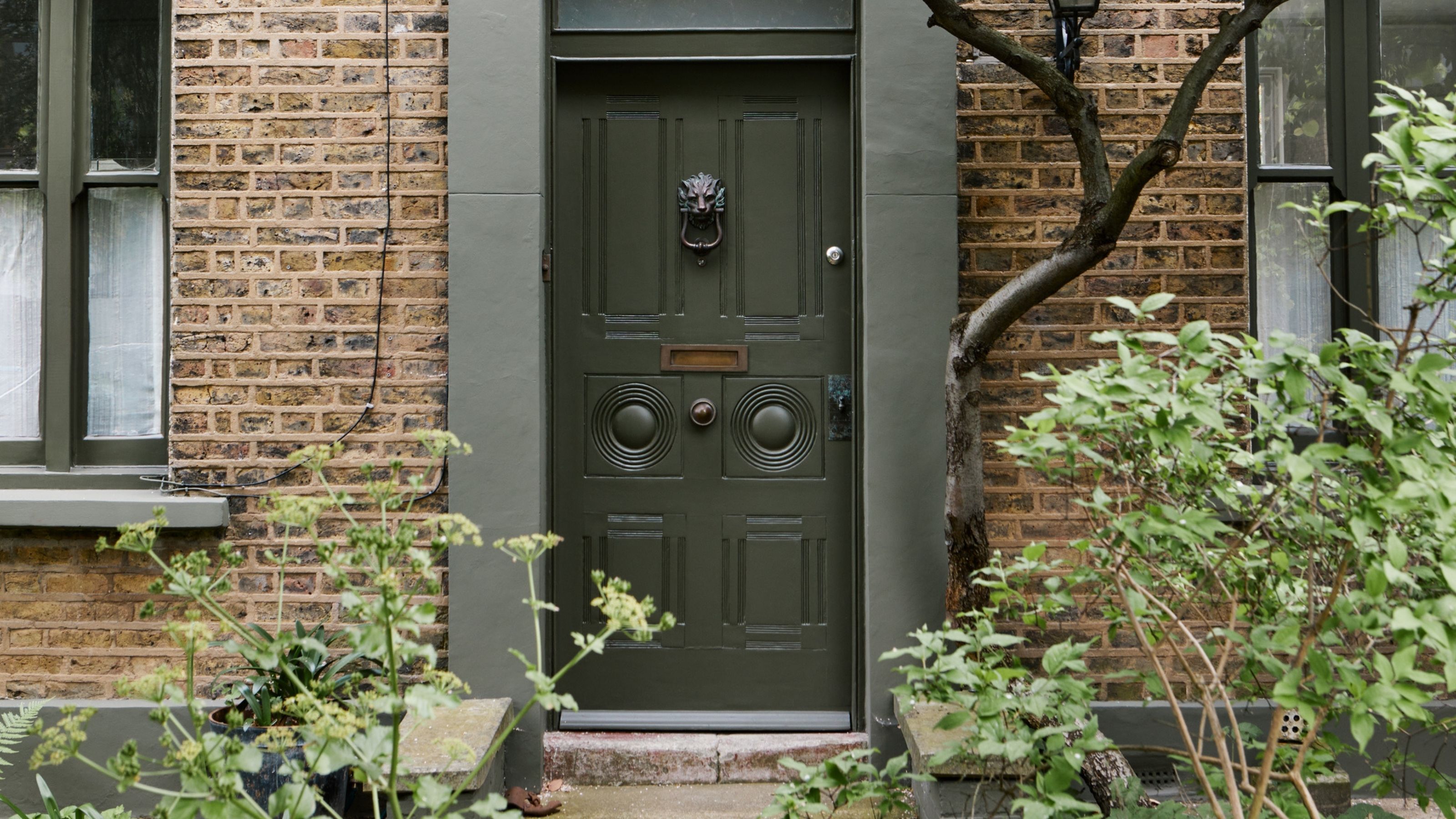 Image of a dark green front door on a light brick home. The window trims and trim around the door are all sage green. There are lots of plants and trees around the house.