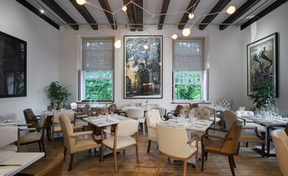 Dining area in the restaurant with leather seating with tables dressed, long white windows, 2 green plants in the corners and walls adorned in art