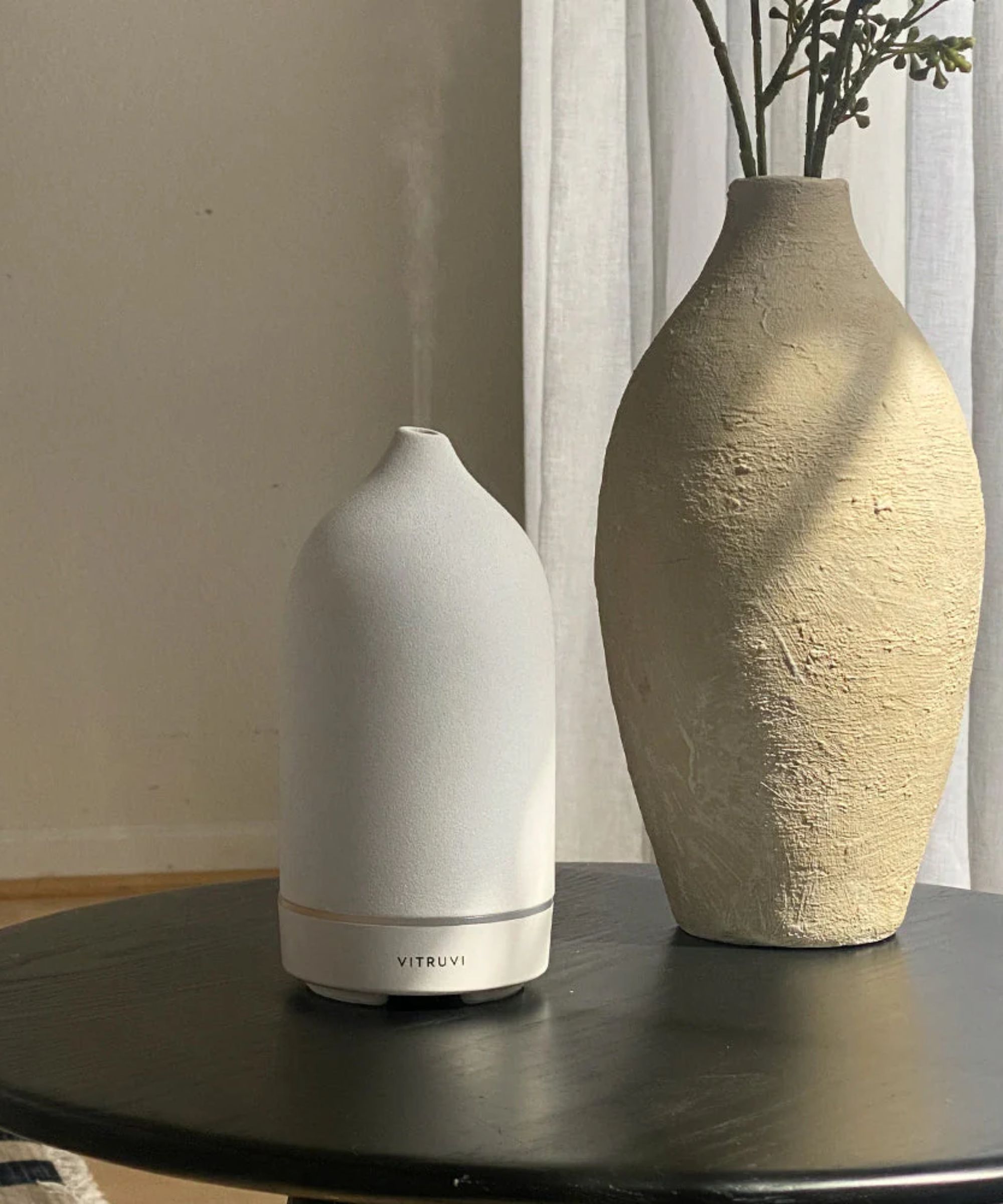A white Vitruvi Stone Diffuser on a black circular table, next to a large beige vase.