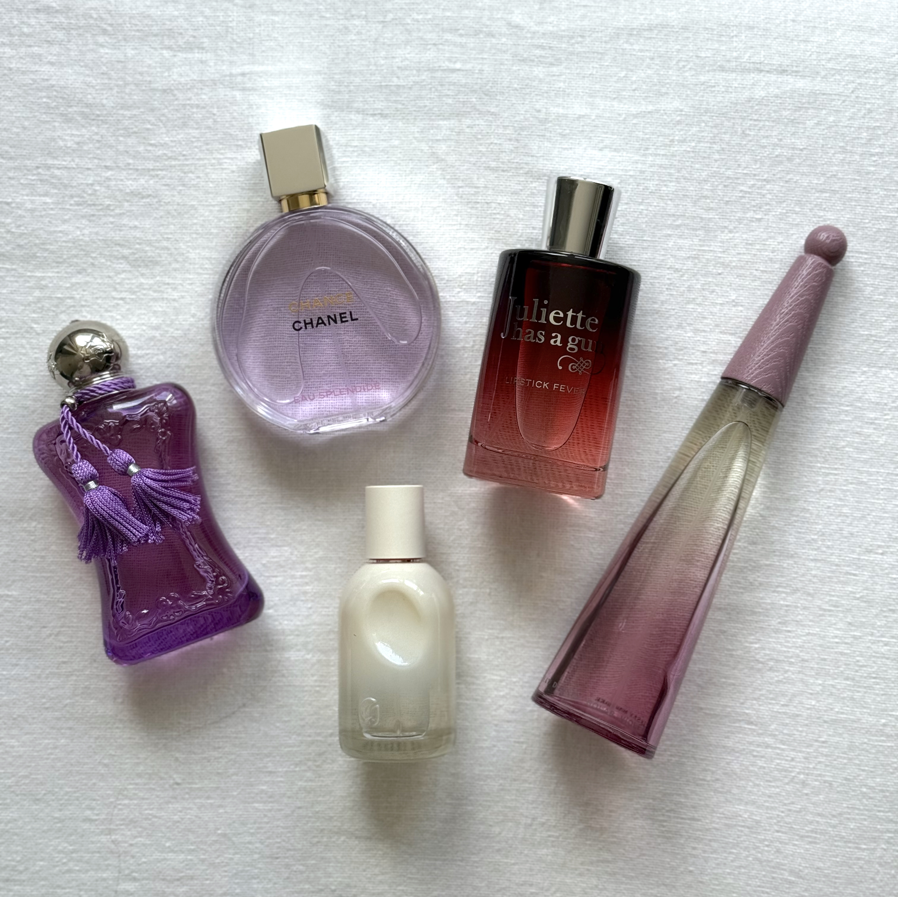 Bird's eye view of a group of violet perfumes from (left to right) Parfums de Marly, Chanel, Glossier, Juliette Has A Gun and Issey Miyake, laying on an off-white linen background