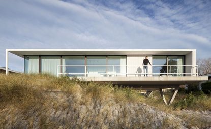 A modern house sits on the Belgian dunes, surrounded by low greenery. A rectangle-shaped house has floor-to-ceiling windows that cover the entire wall. Terra extends over the dunes, and a man is standing on it.