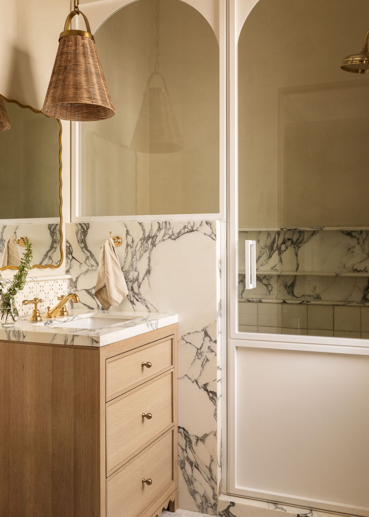 A conservatory style shower in a modern neutral bathroom with white arched frame detailing and marble next to a single vanity unit with a mirror and woven pendant light above it