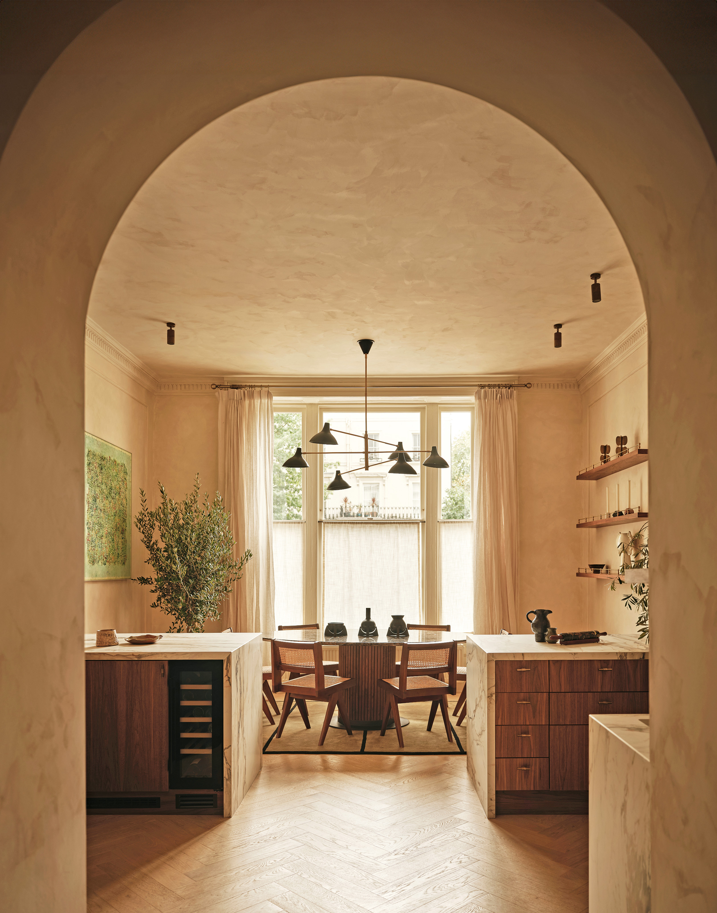 View from arched doorway into kitchen with wood cabinetry, marble tops, wood table and chairs, and layered pendant light