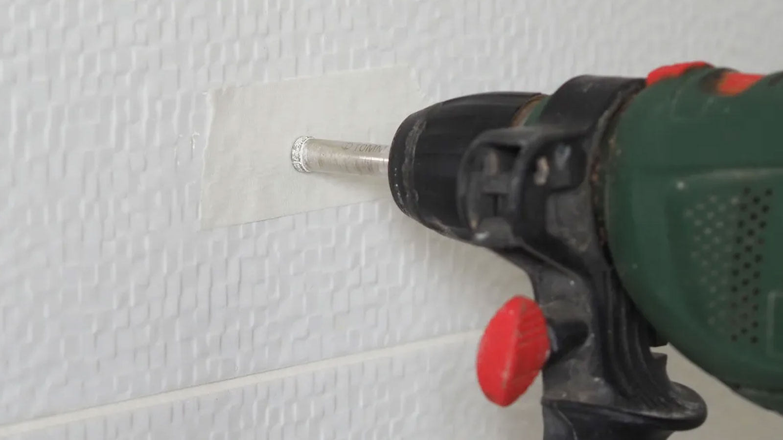 Green drill with diamond tipped drill bit drilling into white tile with masking tape on it