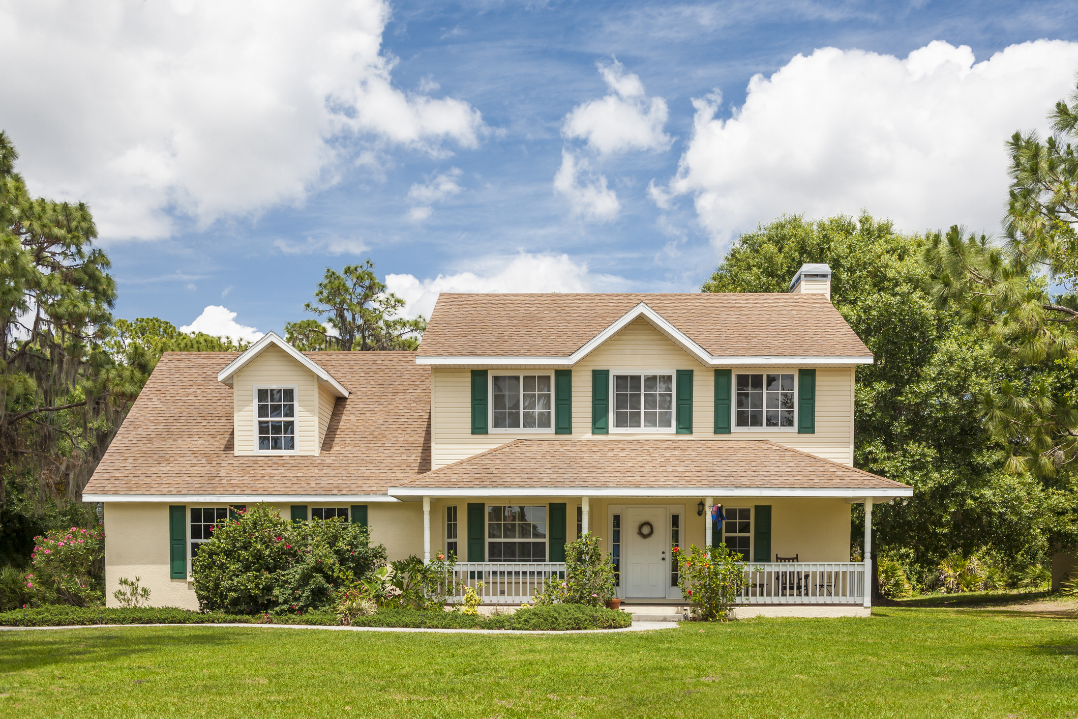 Traditional house in rural setting with porch, decorative shutters, front lawn, flowers and trees. 