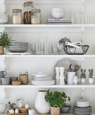 White open shelving with a selection of nested grey and white tableware, glassware, easter linked ornaments such as knitted bunnies and eggs, and potted herbs
