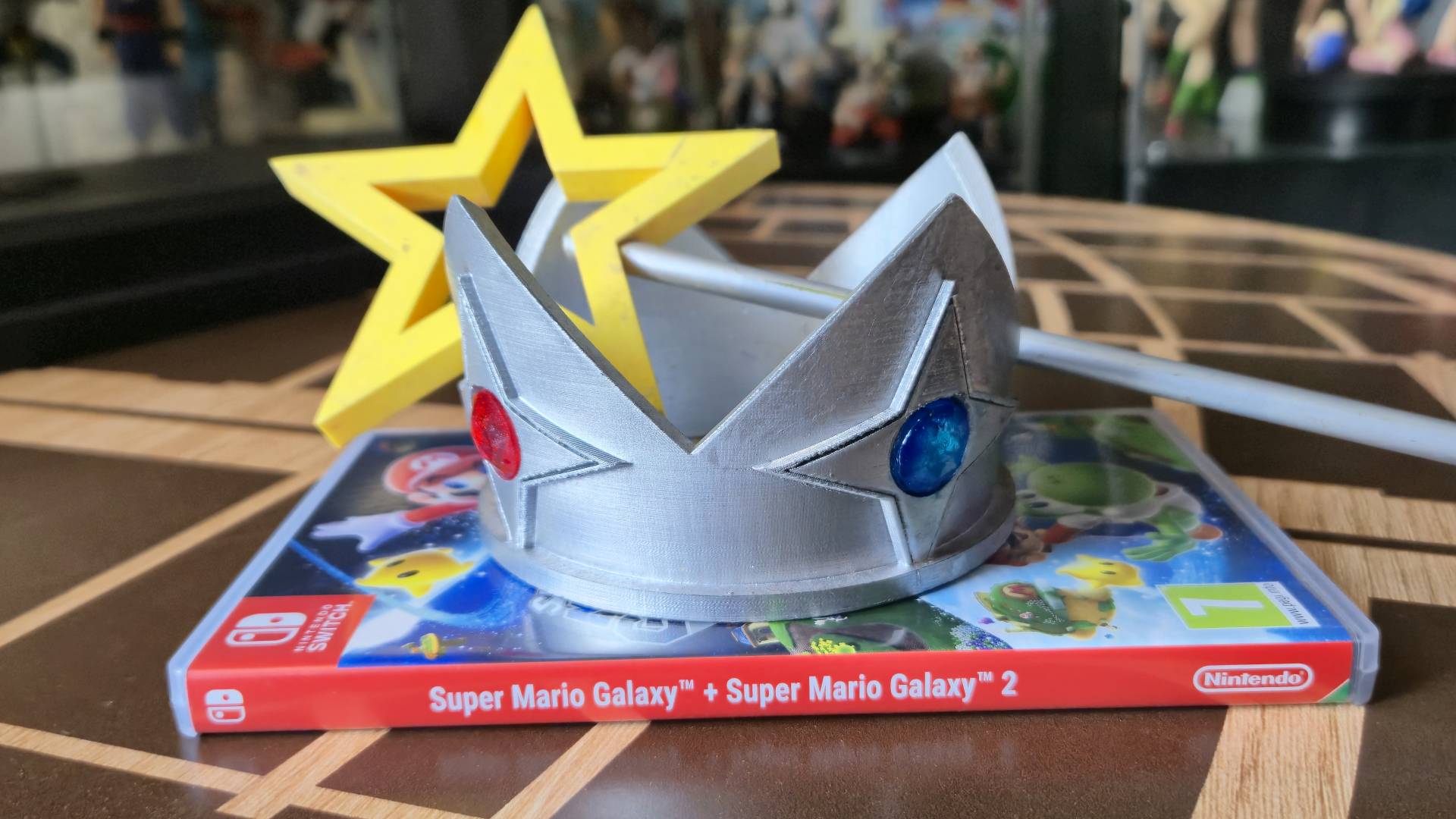 Photo of a physical copy of Super Mario Galaxy + Super Mario Galaxy 2 sitting on a coffee table, with a replica of Rosalina&#039;s crown and wand sitting on top.