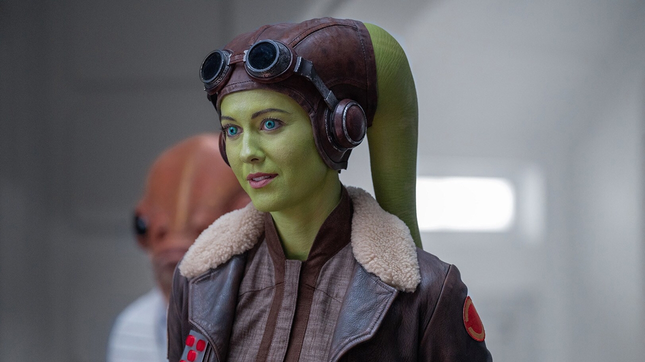 Hera Syndulla is a green-skinned Twi'lek with two head tails (known as lekku) protruding from the top left and right of her head. She is wearing a brown leather cap, goggles, and a brown leather aviator jacket with a fluffy collar. Just behind her is a red squid-like humanoid creature with two large black eyes.