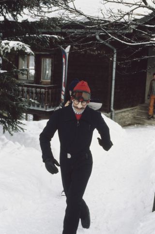 King Charles wearing a black ski suit walking through the snow with fake glasses, a fake mustache and comedy nose