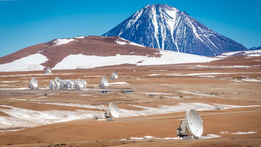 A series of large white radio dishes dot the brown desert with some patches of snow while a large purple mountain looms in the background