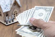 A hand holding cash in front of a new house with keys