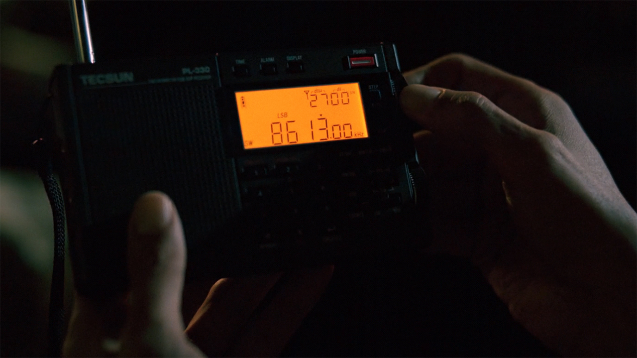 Manousos holding a radio displaying the frequency 8613.00 on it in Pluribus episode 9