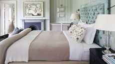 A green bedroom with a double bed with blue velvet headboard, striped wallpaper, artwork above a black fireplace, and a black bedside table with clear lamp with white lampshade on it