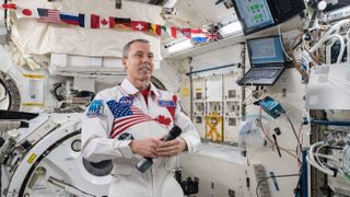 drew feustel floating in a large space station module. he wears a white suit with an american flag across the right half of his chest and a canadian flag across the left half. he faces a camera (not visible in the photo). in the background is a row of flags from different countries. to the right of feustel are two laptops on the wall