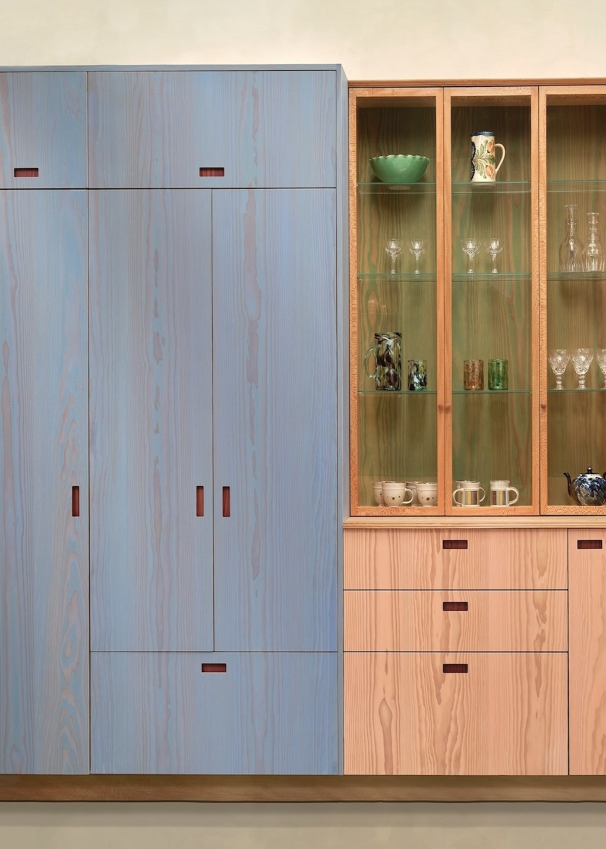 A light blue and salmon painted kitchen cabinets with one side that has glass fronted cabinets. The glass fronted cabinets holds different forms of glassware.