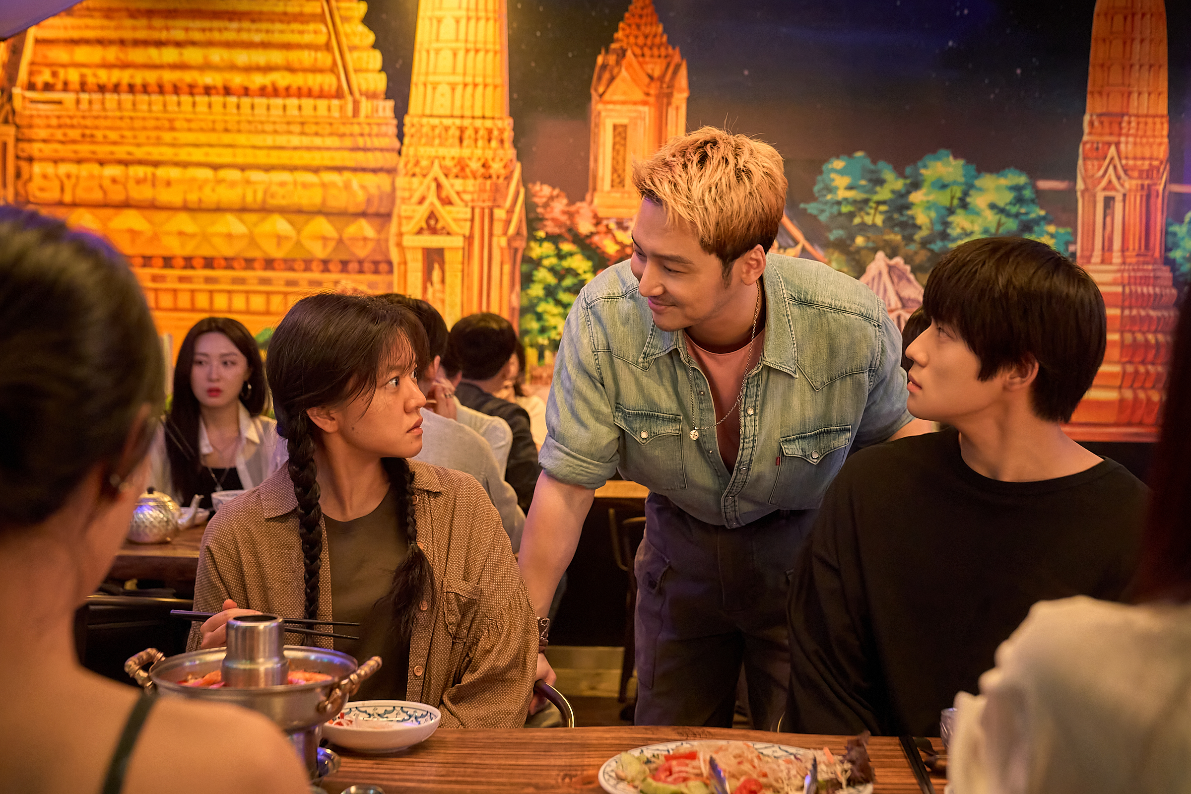 A man with blond hair (Byun Yo-han as Park Yo-han) stands between a teenage girl&amp;nbsp;(Ko A-sung as Kim Mi-jeong) and a teenage boy (Moon Sang-min as Lee Gyeong-rok) as they sit in a crowded Korean restaurant. A painted wall mural is seen behind them, in a still from the Korean romance movie 'Pavane.'