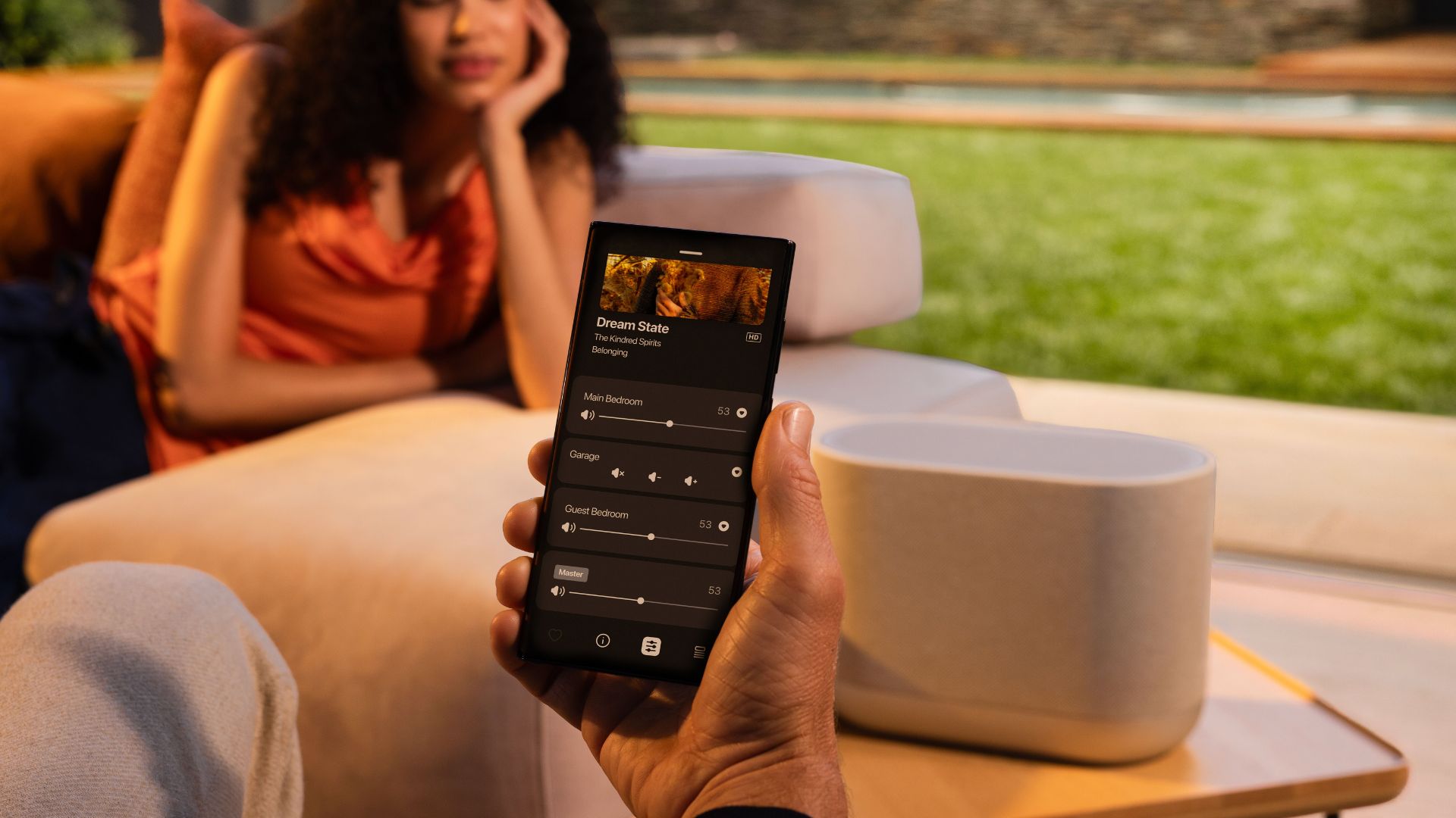 A close up of a smartphone running the HEOS app with a Denon home speaker in the background