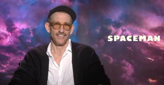 a man in a hat and glasses sits in front of a graphic of a spaceship flying through purple clouds