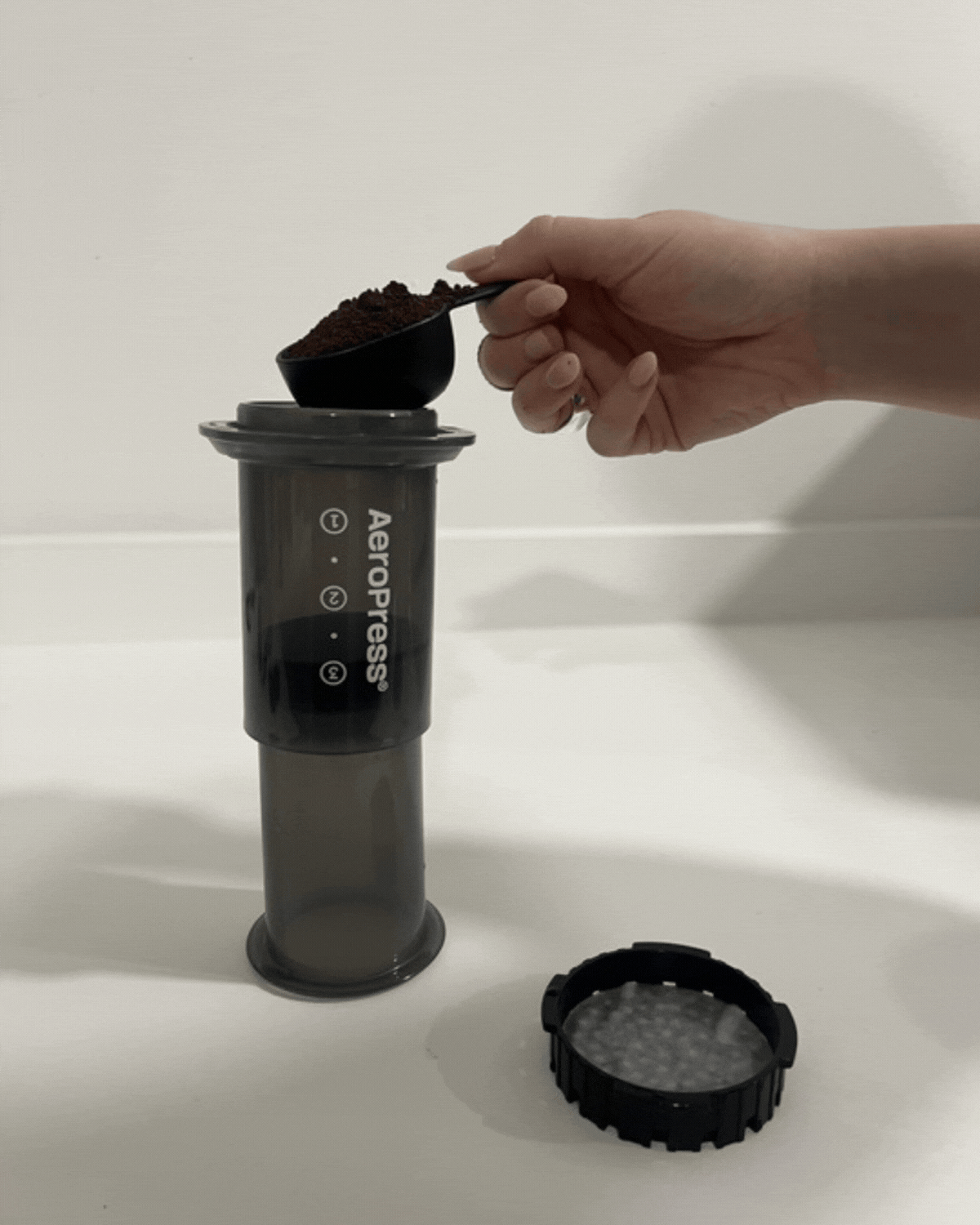 GIF of a hand dumping a scoop of coffee grounds into a black AeroPress coffee maker.