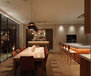 A contemporary kitchen with a small island featuring a white marble waterfall countertop and two blue velvet stools with gold legs. Overhead, two sculptural pendant lights with brass detailing and frosted glass globes add elegance. The cabinetry combines textured bronze and wood finishes, complemented by marble backsplash and under-cabinet lighting.