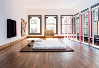 The bedroom has six large windows with dark blue window frames. There is a mattress in the center of the room, set on a wooden platform.