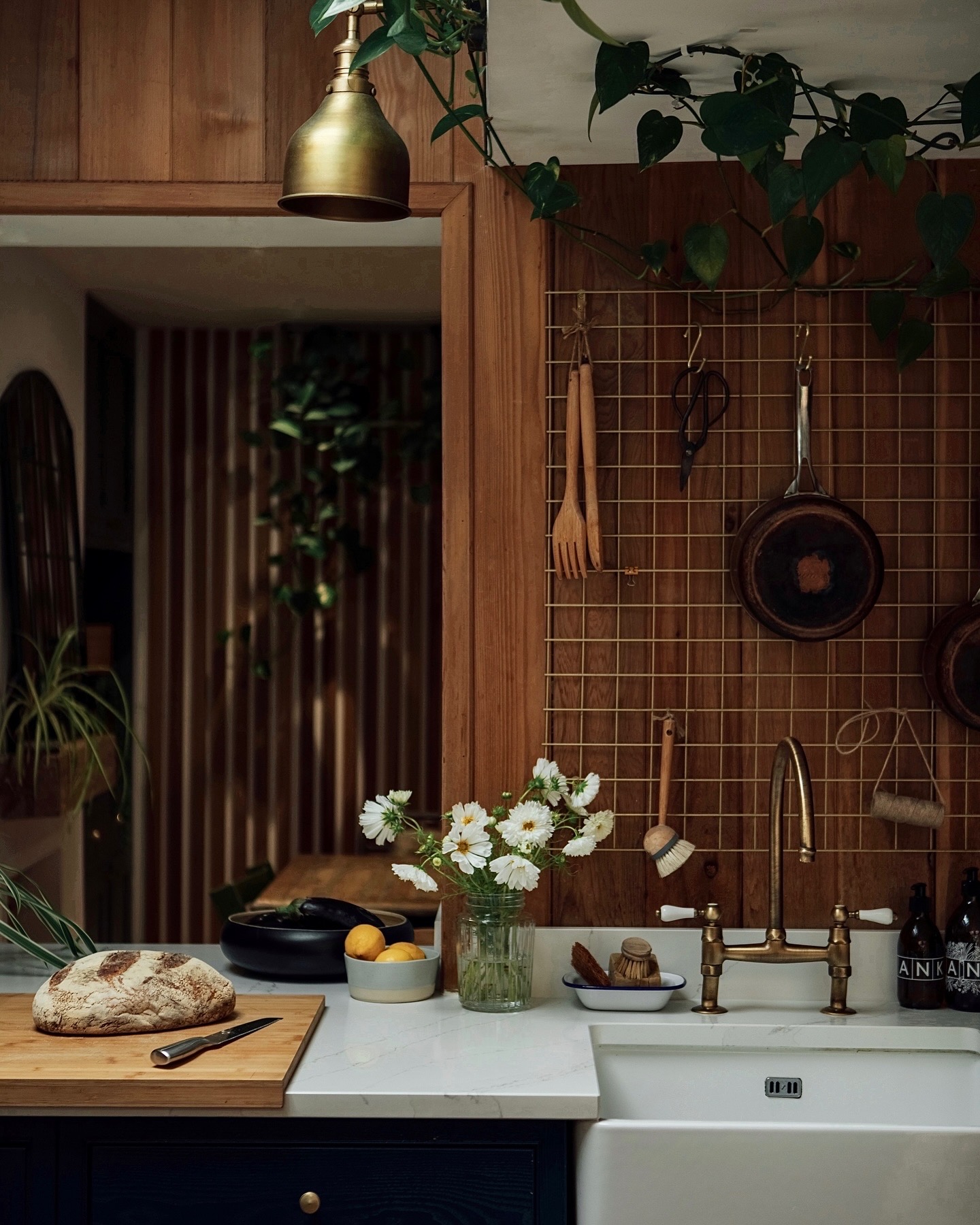 a close up on a kitchen with light stone counter, cutting board with a loaf of bread, glass with flowers, sink with brass tap, wire screen with utensils hanging off it