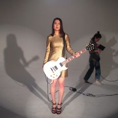 mitski stands holding a bass guitar in a gold dress on the set of her music video your best american girl