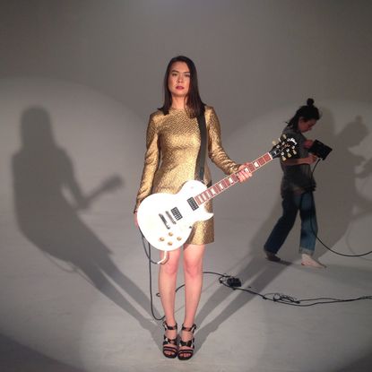 mitski stands holding a bass guitar in a gold dress on the set of her music video your best american girl