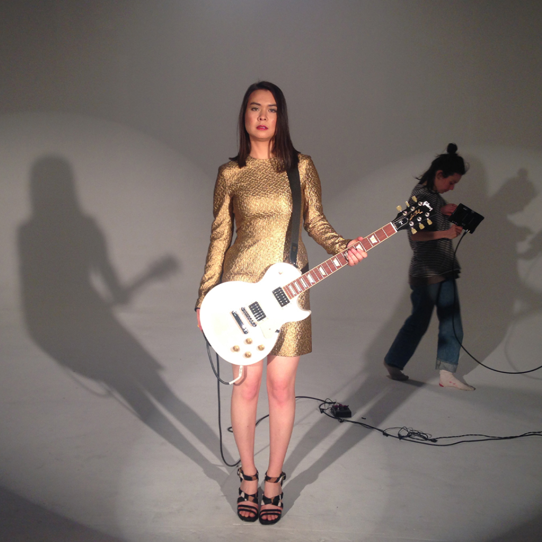 mitski stands holding a bass guitar in a gold dress on the set of her music video your best american girl