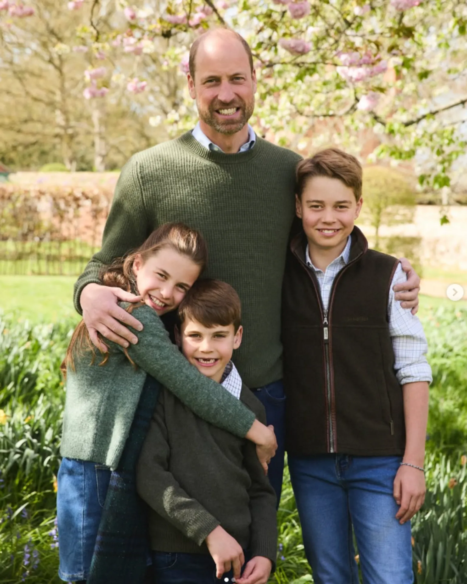 Prince William wearing a green sweater standing with his arms around Prince George, Princess Charlotte and Prince Louis in a garden