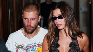 Justin and Hailey Bieber in New York City walking down the street. She has on sunglasses and a black satin gown and he's wearing a graphic tee with cats on it. 