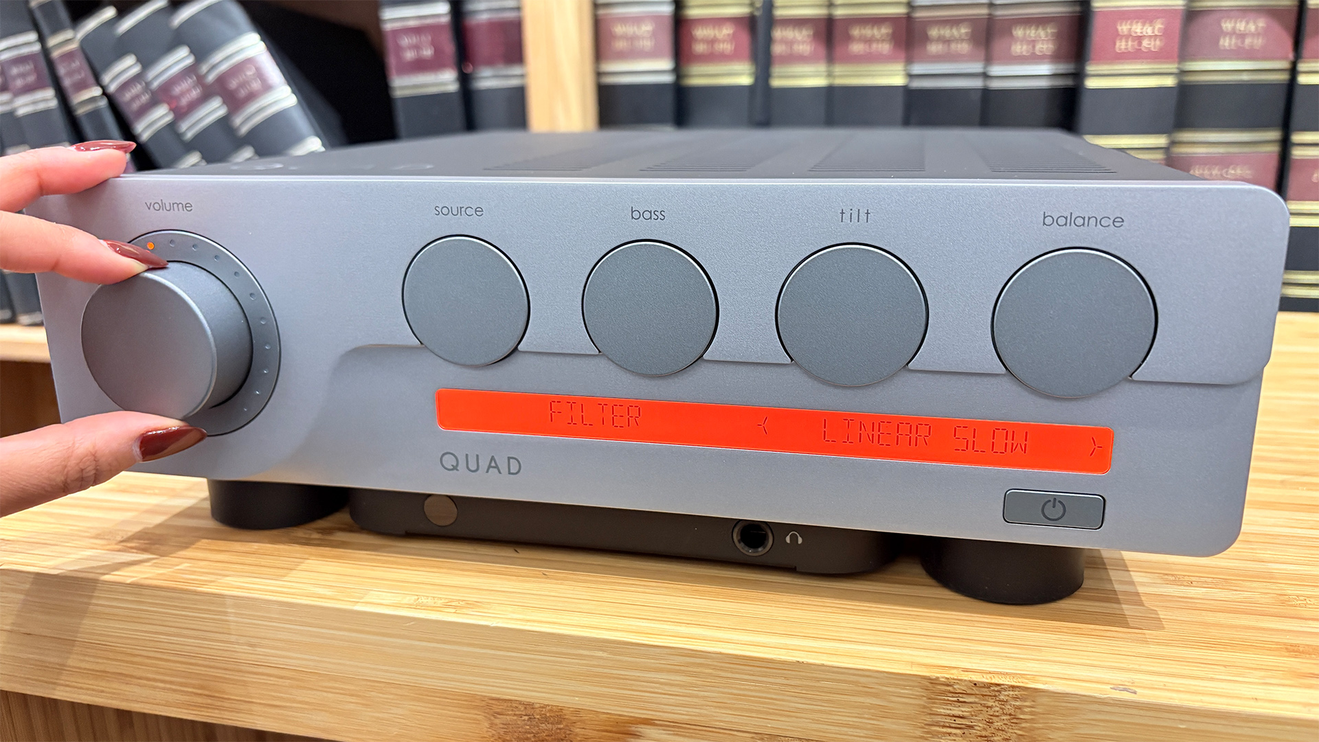 Quad 3 integrated amplifier on wooden hi-fi rack in front of bookcase, hand is visible adjusting left control dial