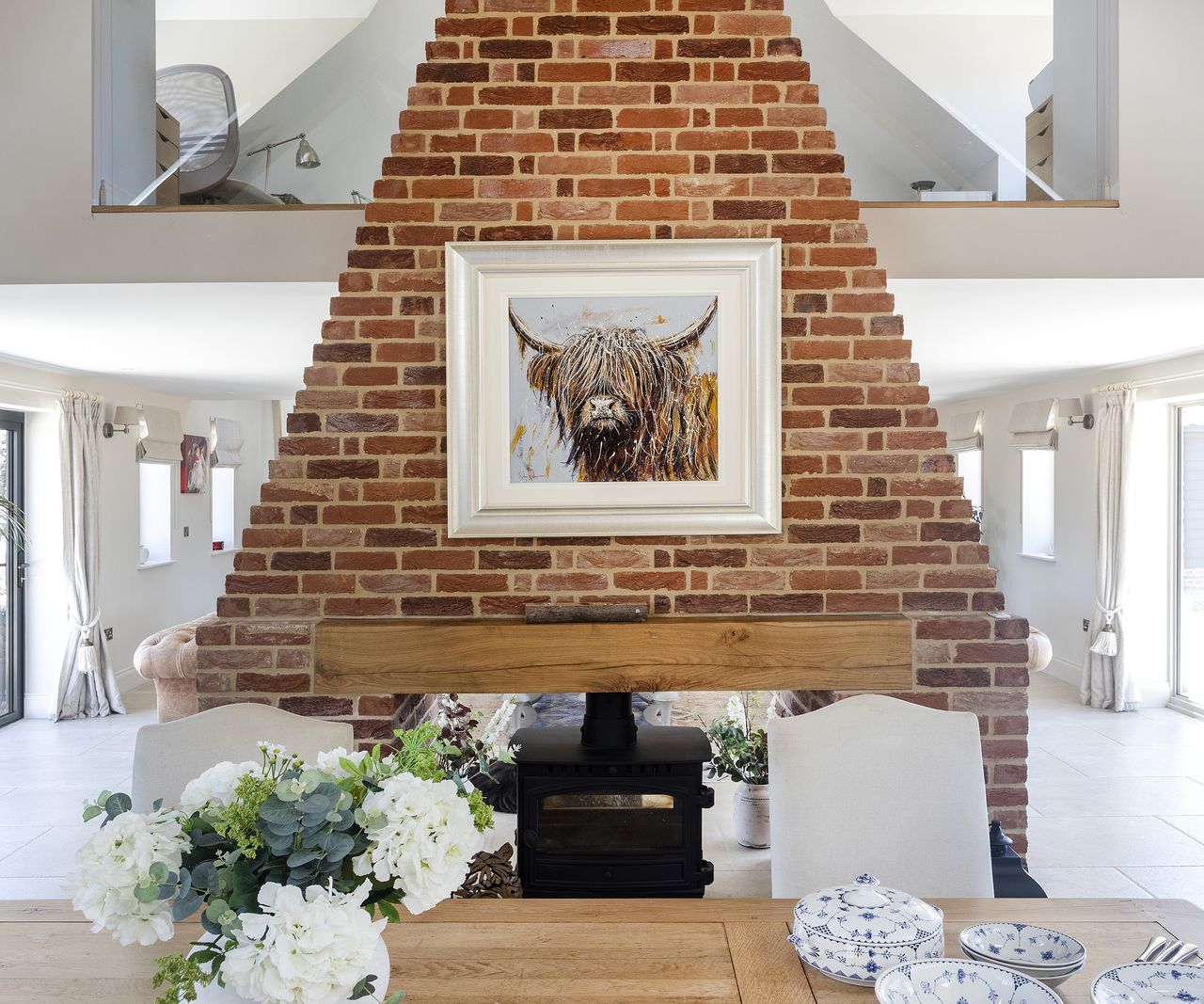 A red brick chimney built in the centre of a large downstairs around a log burning stove with a mezzanine level in the background