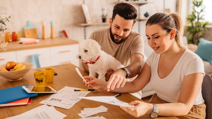 A young couple go over paperwork at their kitchen table while their dog looks on.