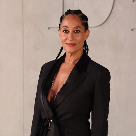 a black woman wears a black blazer and cornrows while posing in front of a white wall