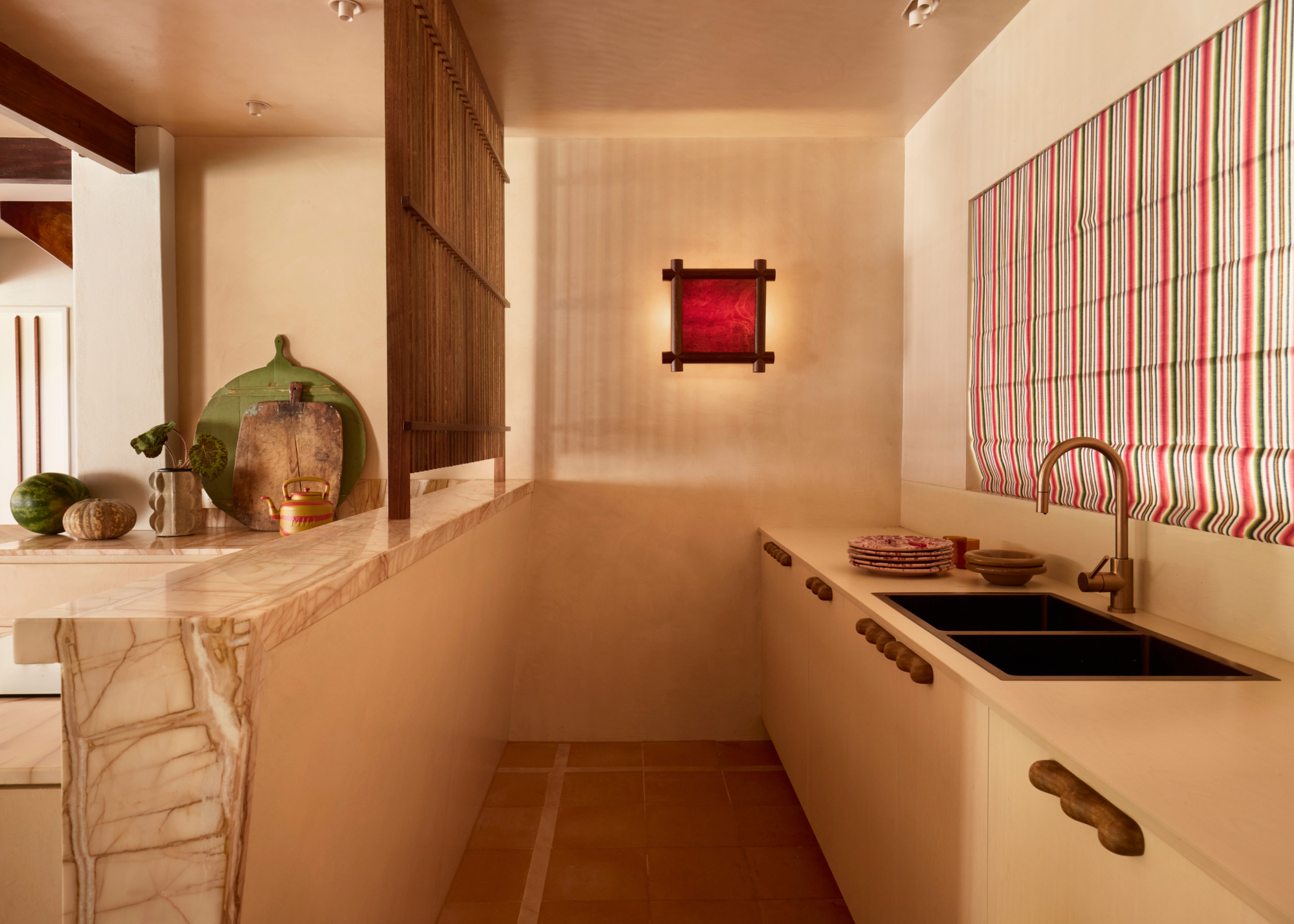 A modern compact kitchen area in neutral tones with an abstract wall light that looks like artwork, striped closed blinds above the sink and a marble-topped peninsula
